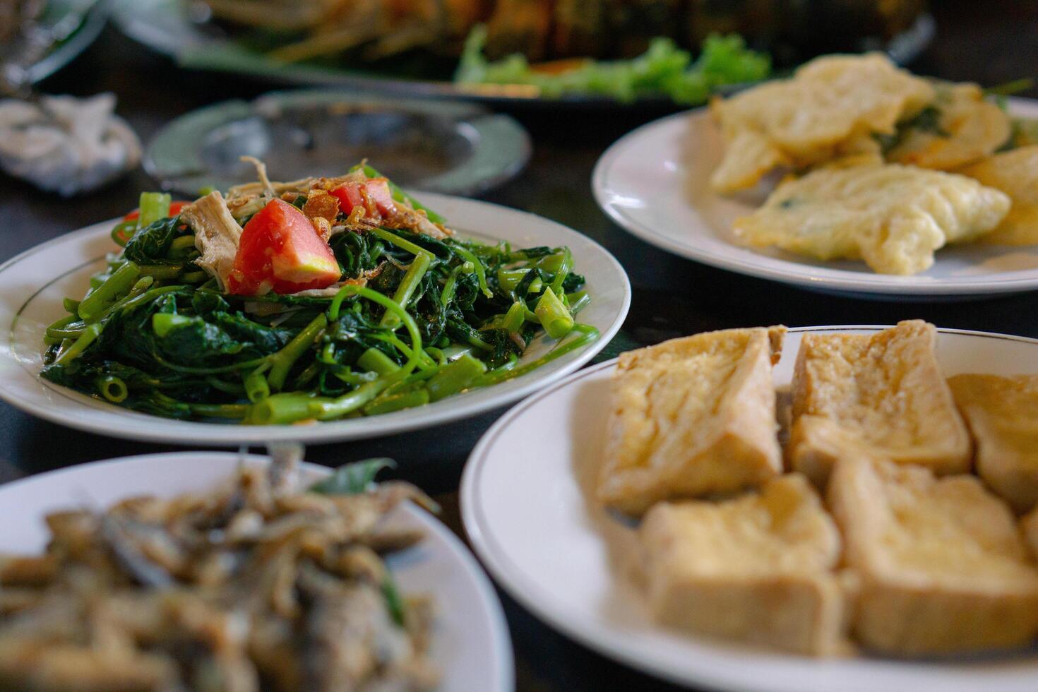 Cah kangkung or Stir fried water spinach is ready to eat on white plate. Surrounded by fried fish and fried tofu. Asian local cuisine and Indonesian food photo