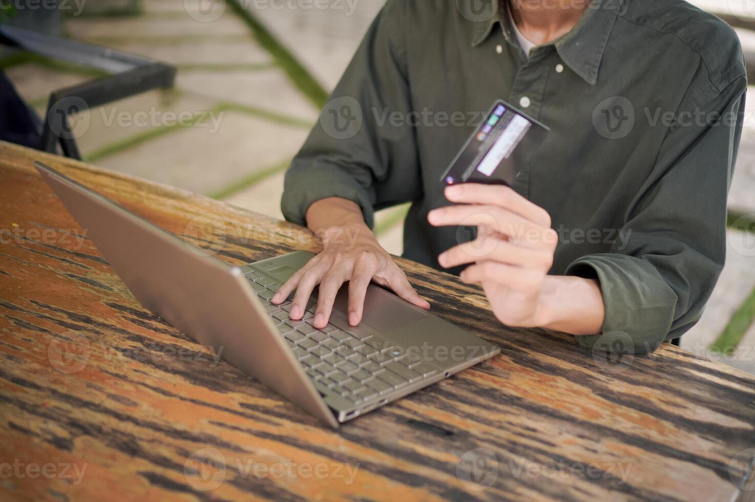 Young Asian man working on his laptop while talking on mobile phone and holding a credit card. Man trying to online purchasing with card photo