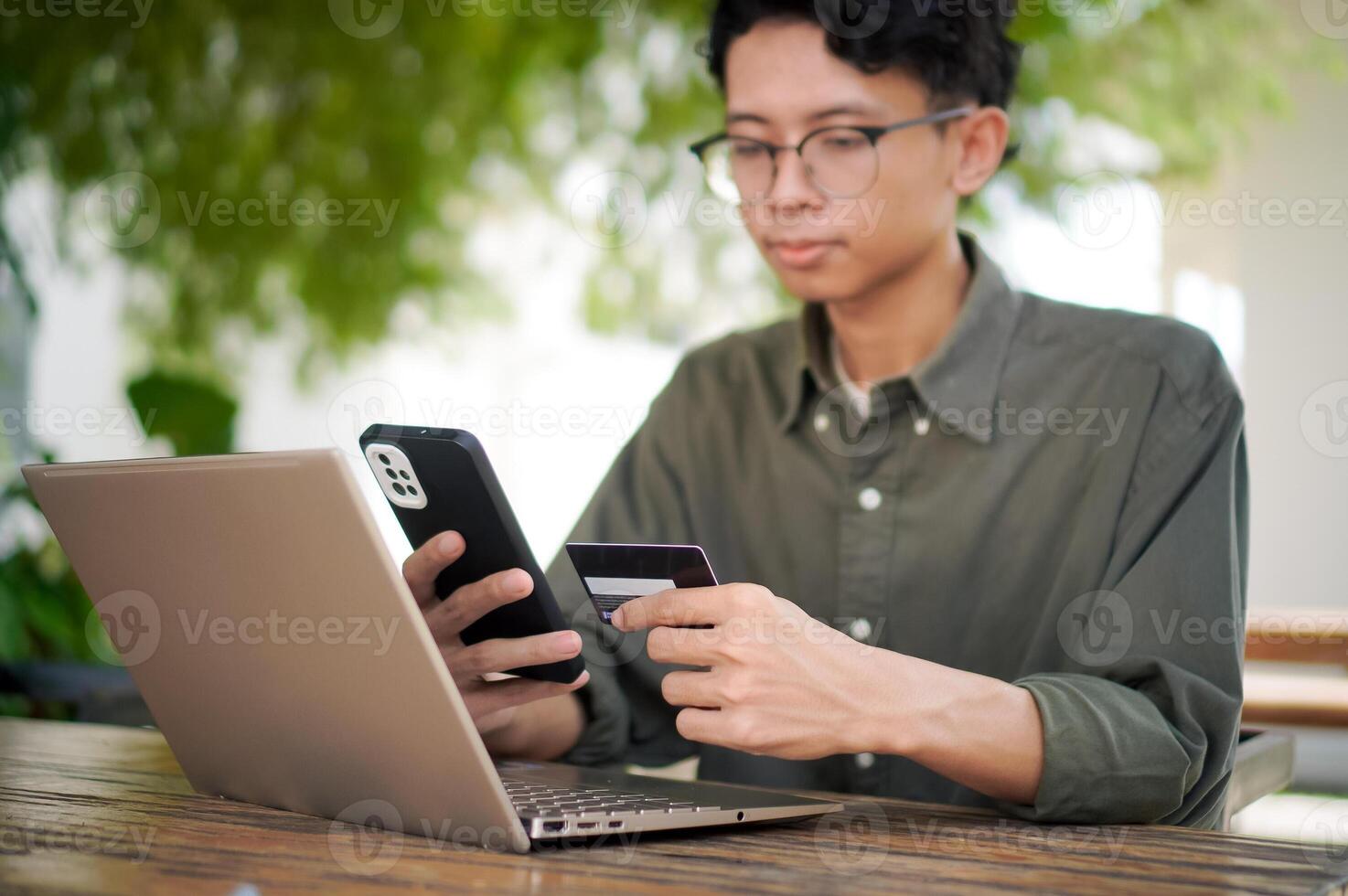 Young Asian man working on his laptop while talking on mobile phone and holding a credit card. Man trying to online purchasing with card photo