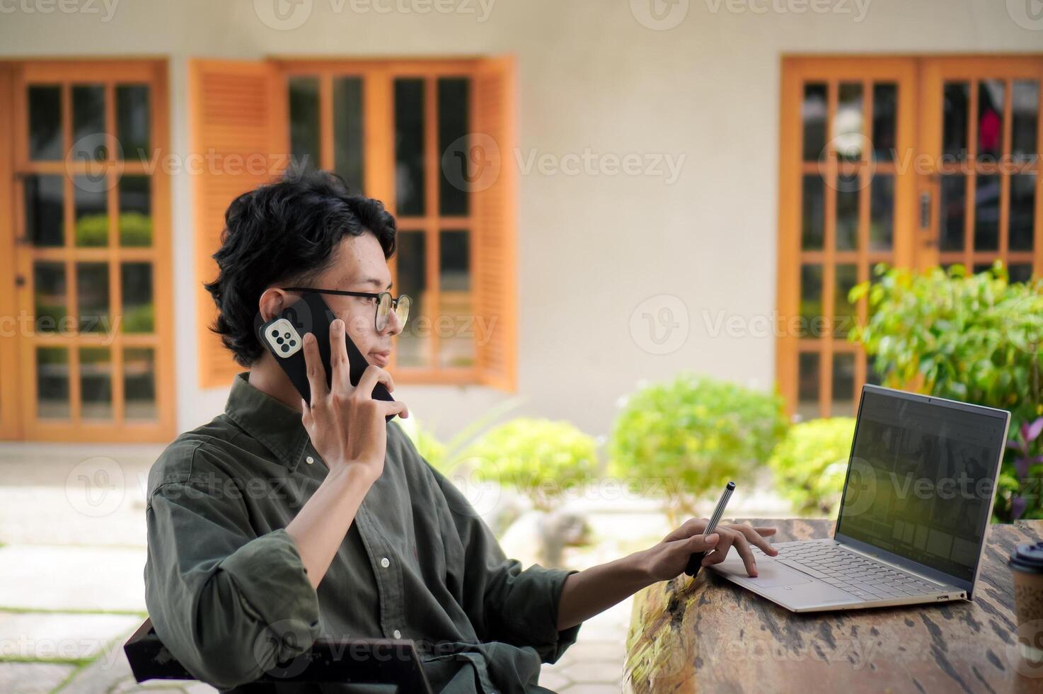 Outdoor shot of young Asian man working on his laptop while talking on mobile phone. photo