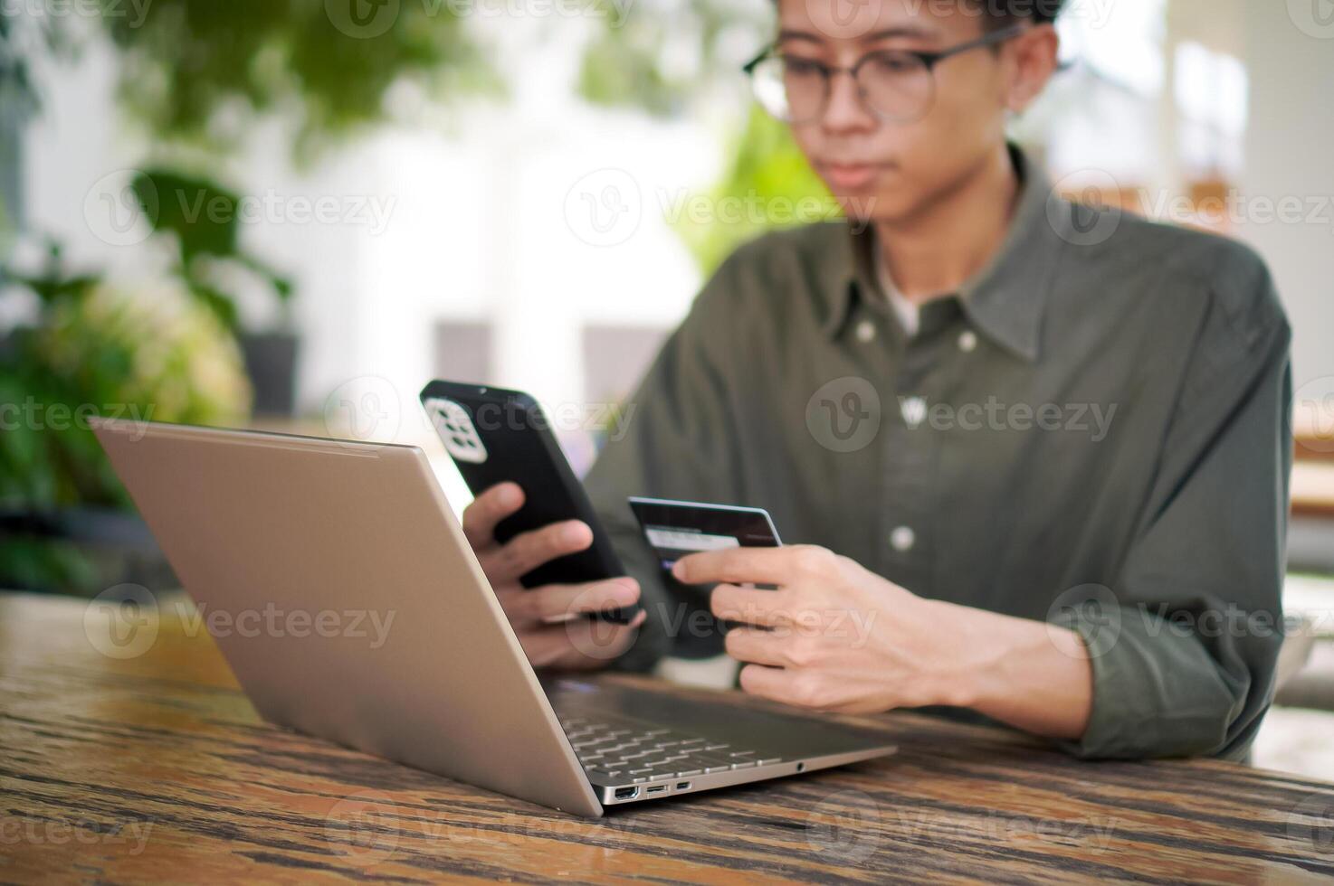 Young Asian man working on his laptop while talking on mobile phone and holding a credit card. Man trying to online purchasing with card photo