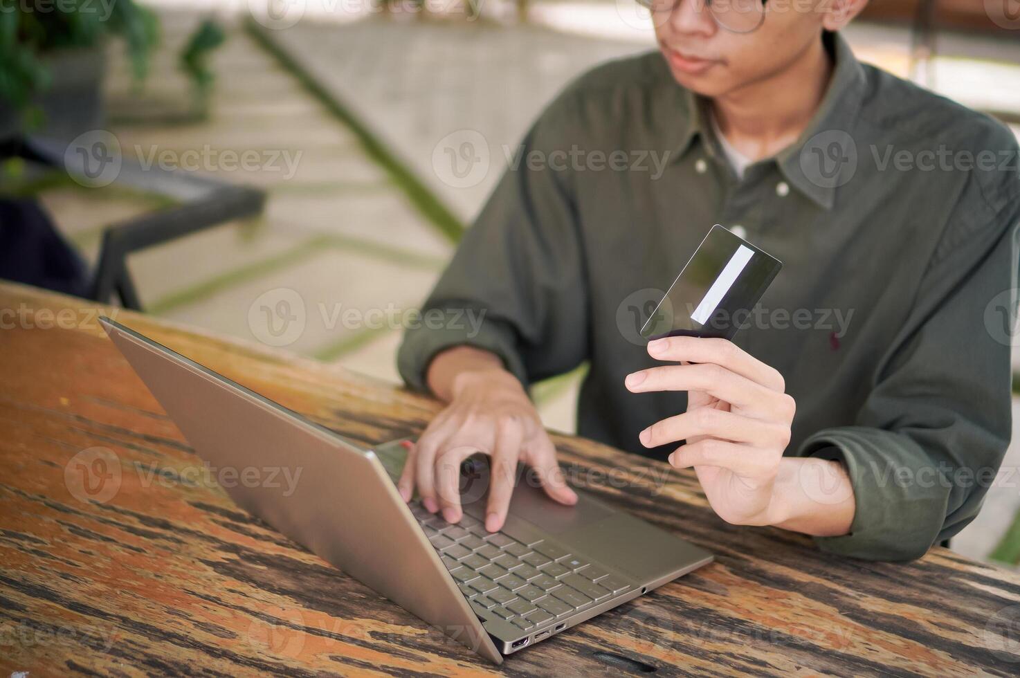 Young Asian man working on his laptop while talking on mobile phone and holding a credit card. Man trying to online purchasing with card photo