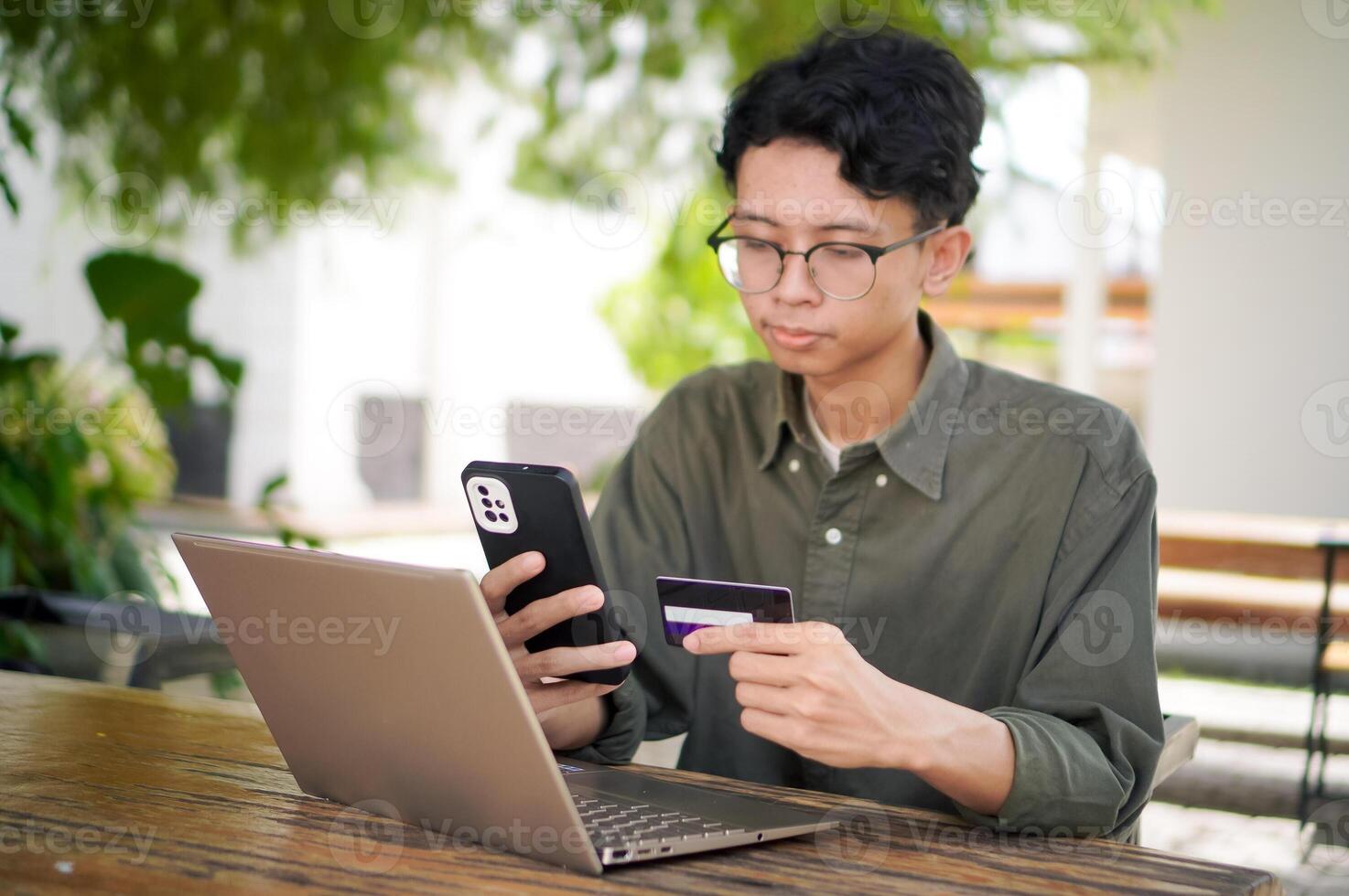 Young Asian man working on his laptop while talking on mobile phone and holding a credit card. Man trying to online purchasing with card photo