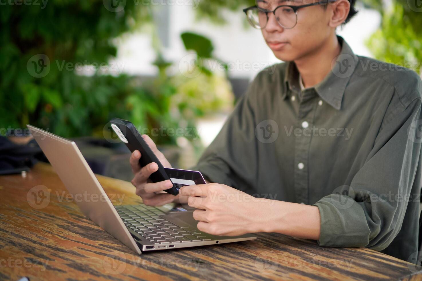 Young Asian man working on his laptop while talking on mobile phone and holding a credit card. Man trying to online purchasing with card photo