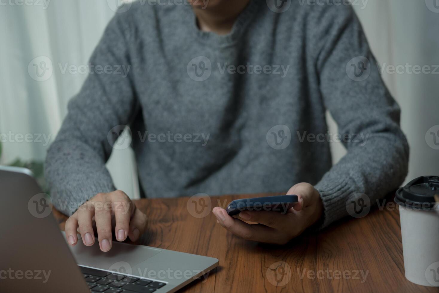 Man in gray sweater using a laptop and holding a smartphone at a wooden table with a coffee cup, focusing on multitasking in a work-from-home setup. photo