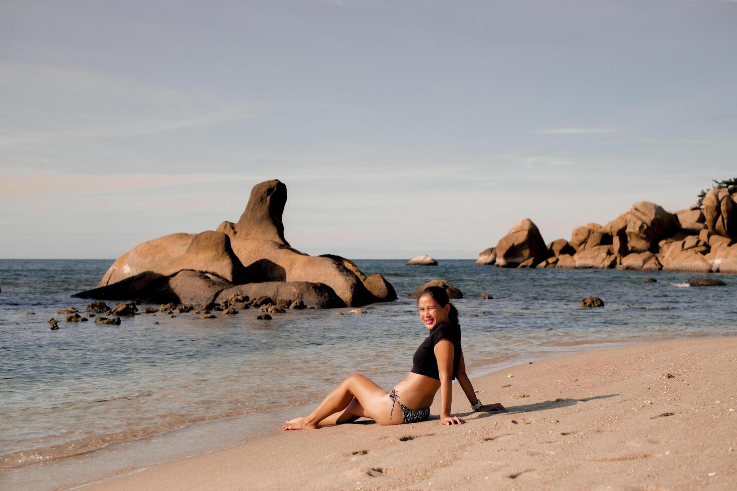 View of Asian woman in black bikini swimsuit sitting on the beach on sunny day, enjoying sunshine. Summer, holiday, travelling, vacation. Woman relax by ocean for summer vacation. Samui, Thailand. photo