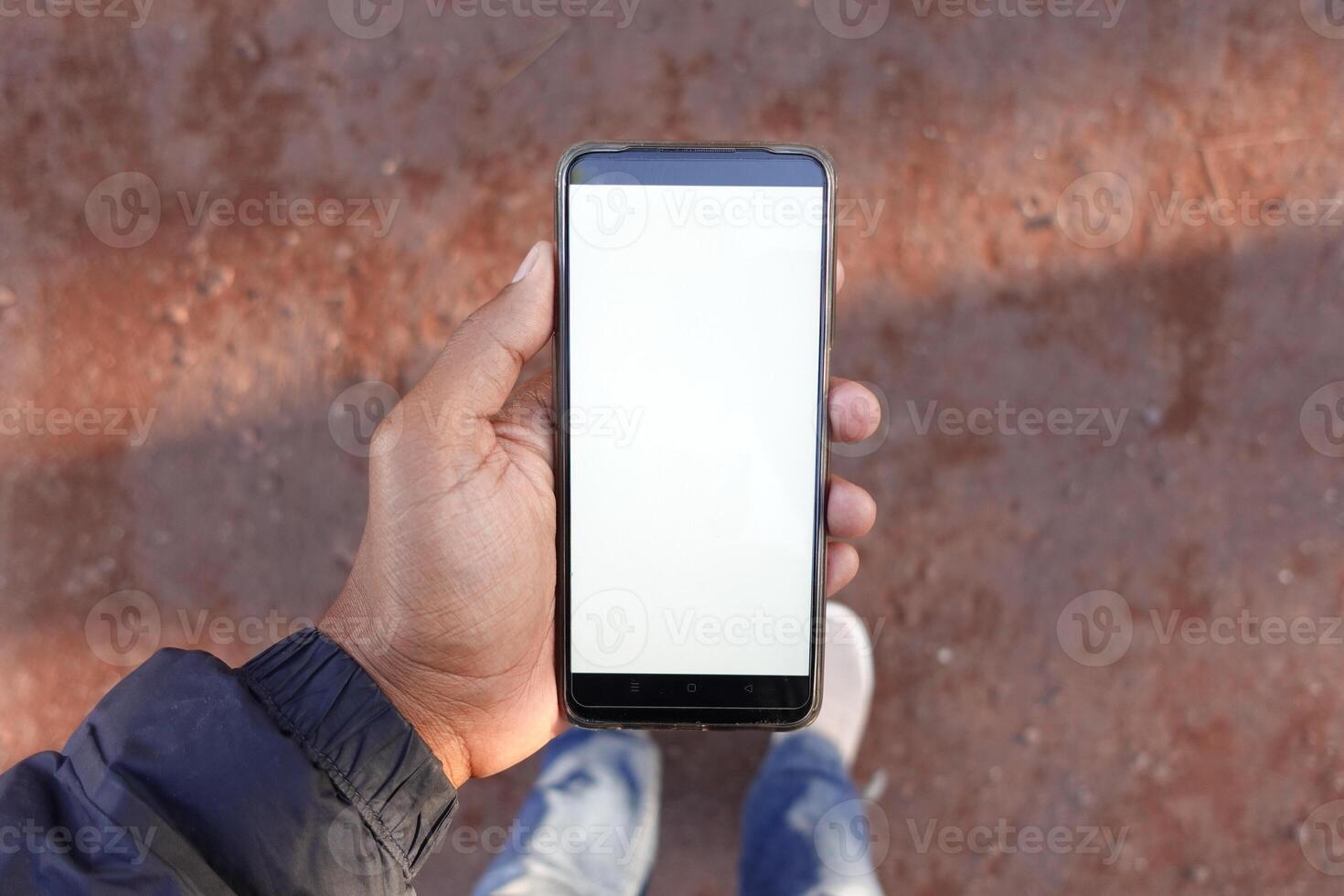 A hand firmly grasping a smartphone that has a blank screen positioned on the ground surface photo