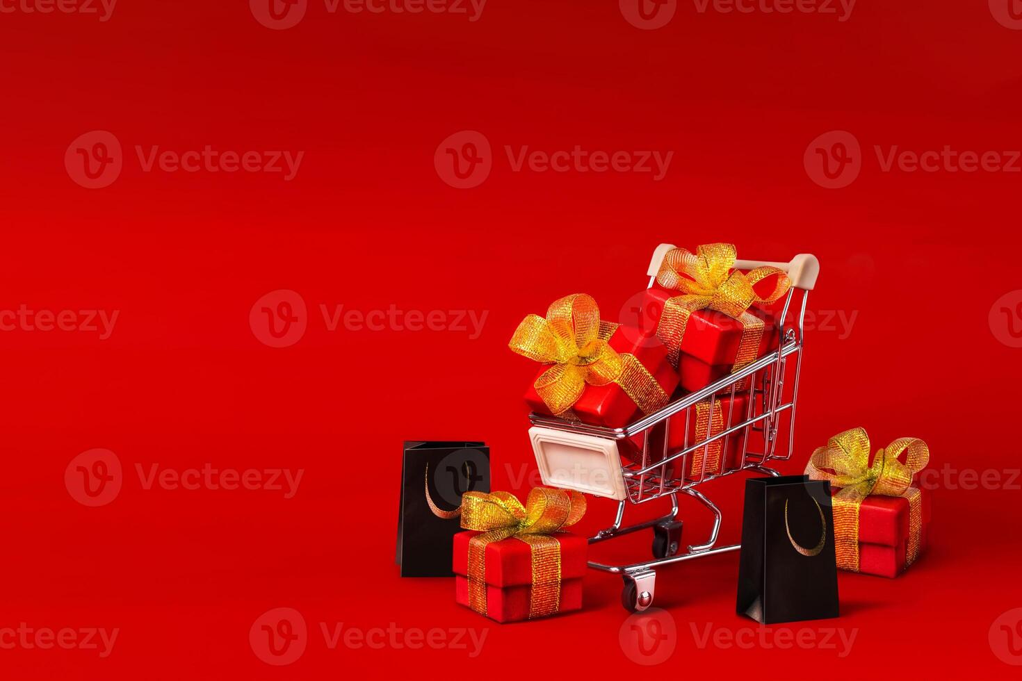 Supermarket trolley with red box with gold bows and black shopping bags. The concept of buying gifts for holidays. Discounts, Black Friday. Red background, Copy space photo