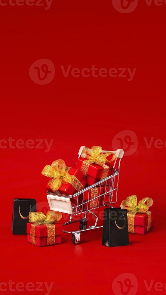 Supermarket trolley with red box with gold bows and black shopping bags. The concept of buying gifts for holidays and sales. Discounts, Black Friday. Red background, Copy space vertical banner photo