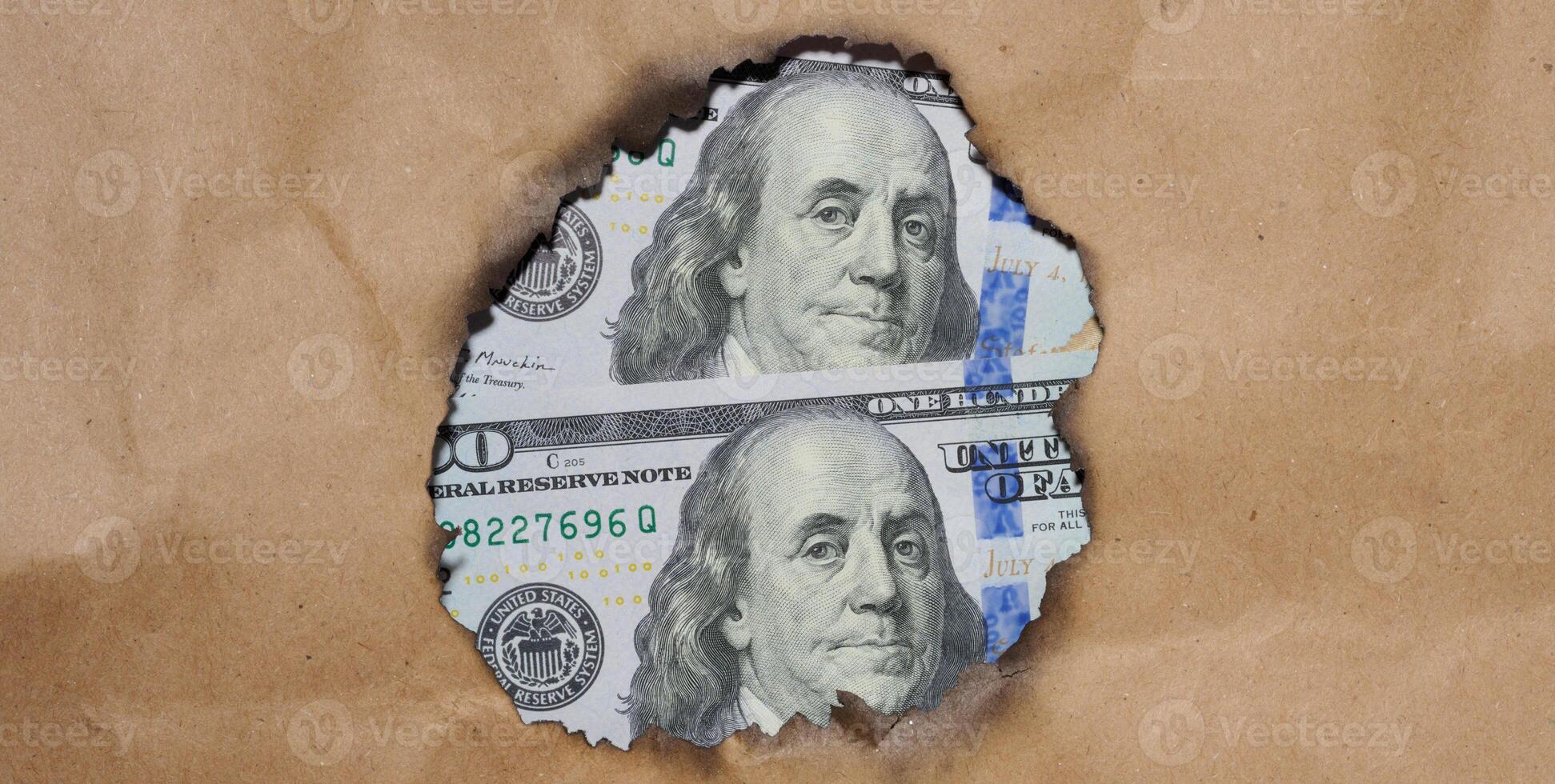A portion of a US bill featuring Benjamin Franklin, the bill visible through a burnt hole in the brown paper photo