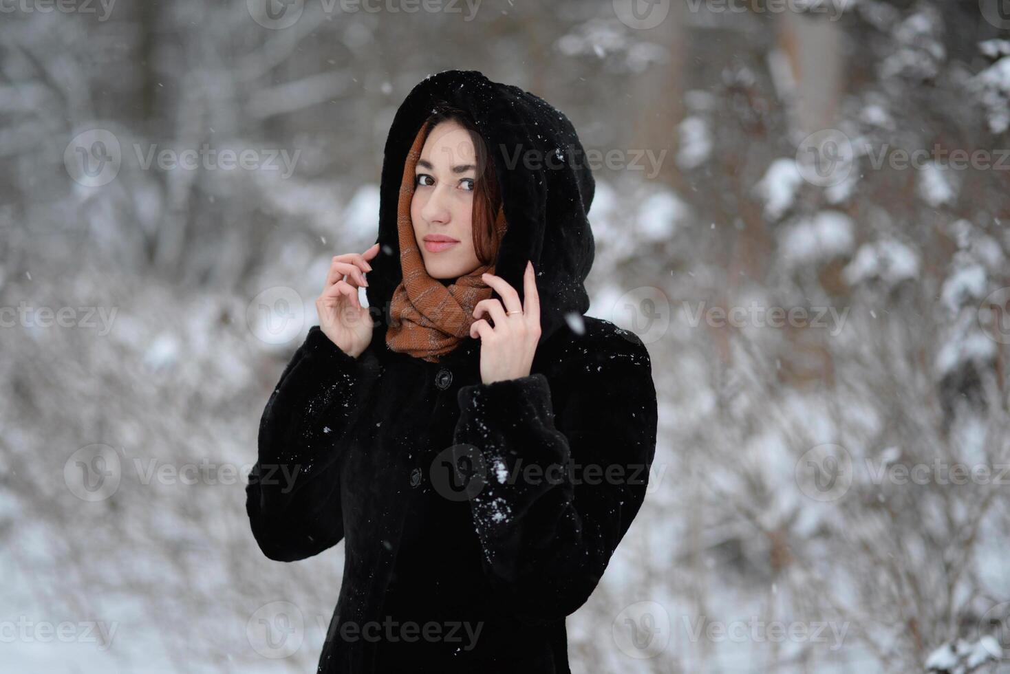 A girl wearing a black coat with a hood posing in a snowy forest in winter. Cold frosty snowy winter weather concept photo