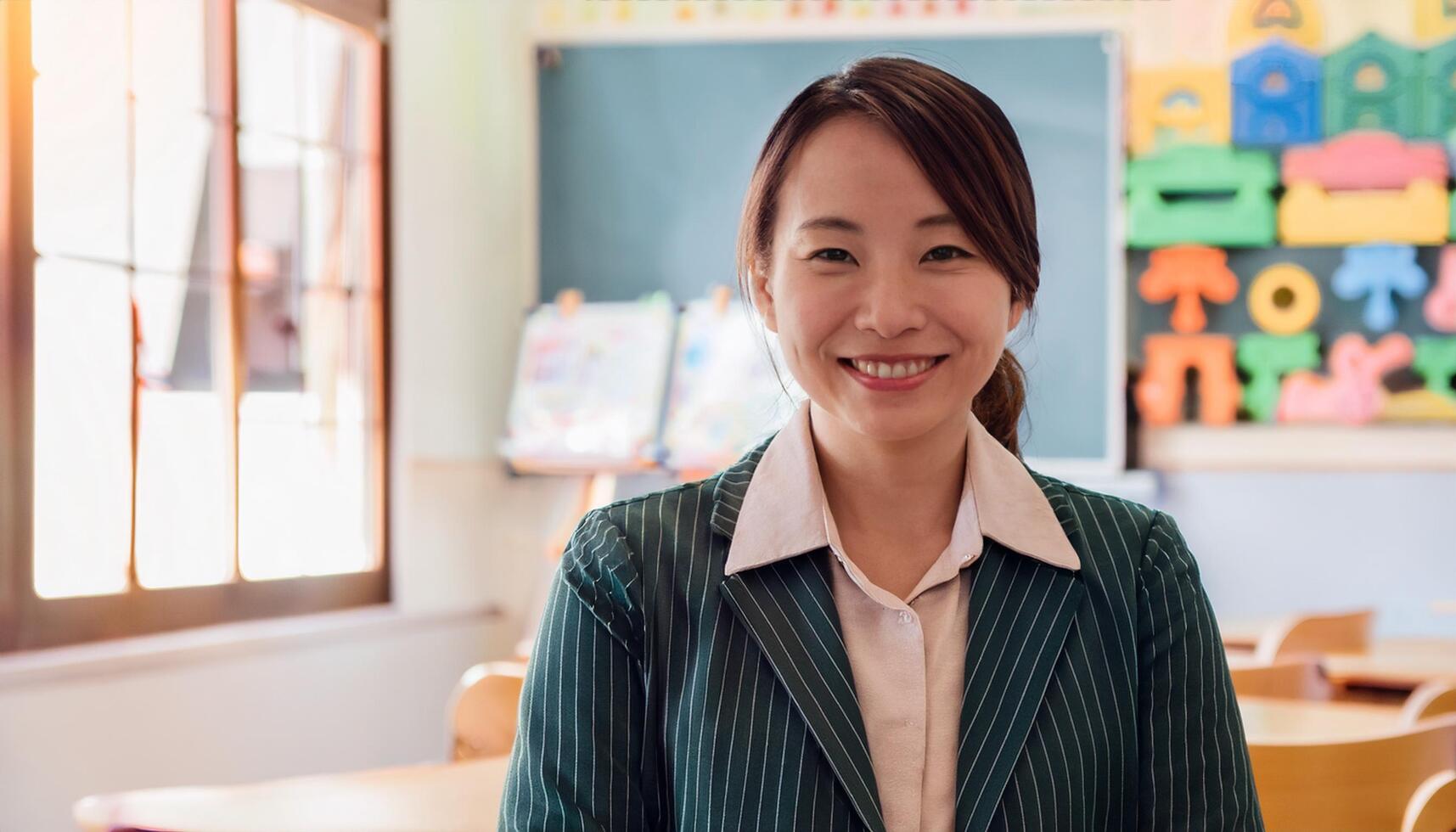 Japanese preschool teacher smiling warmly in a classroom filled with children. The teacher, The vibrant learning environment in the background makes this image perfect for educational themes, school photo
