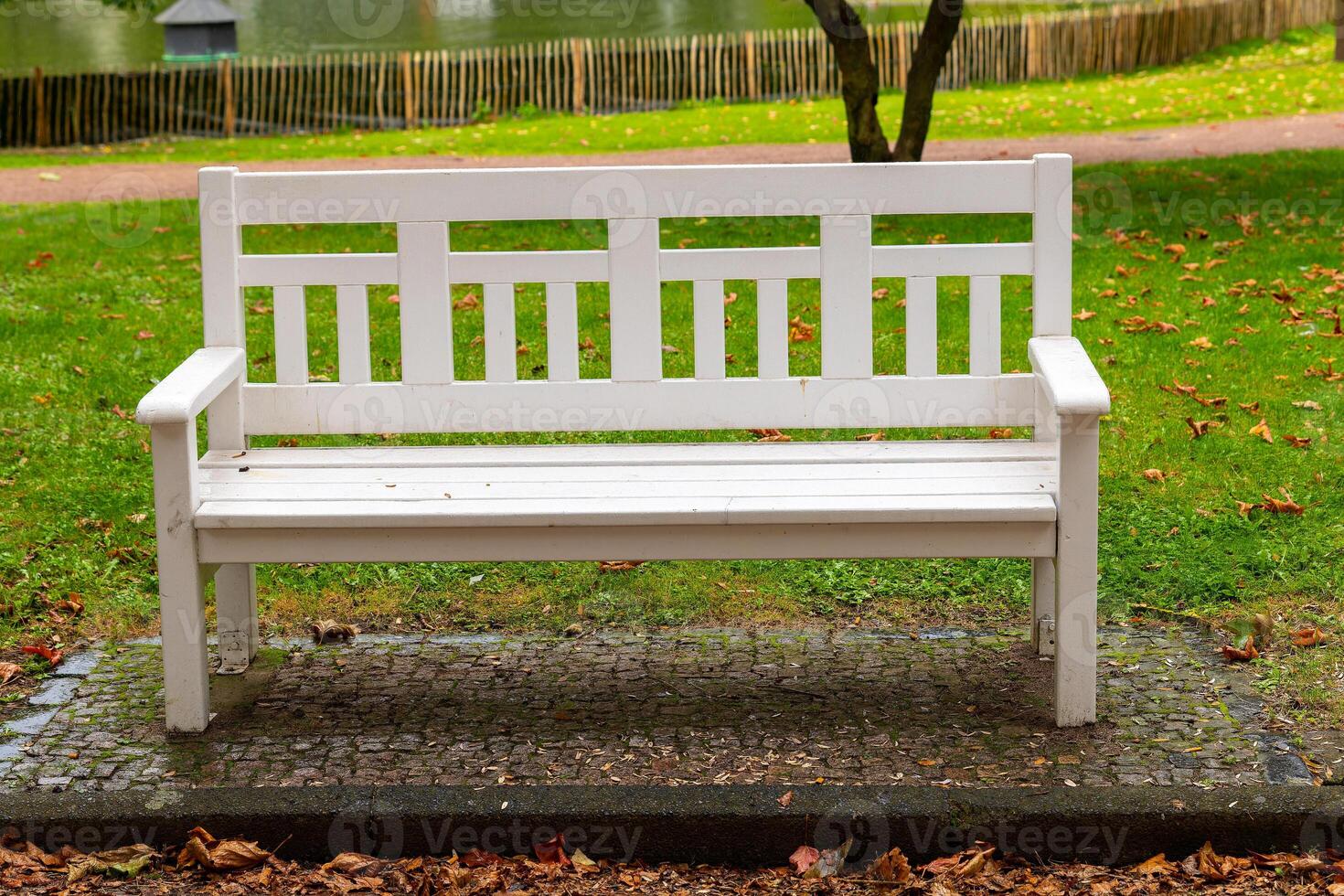 white bench sits in a park with a fence in the background photo