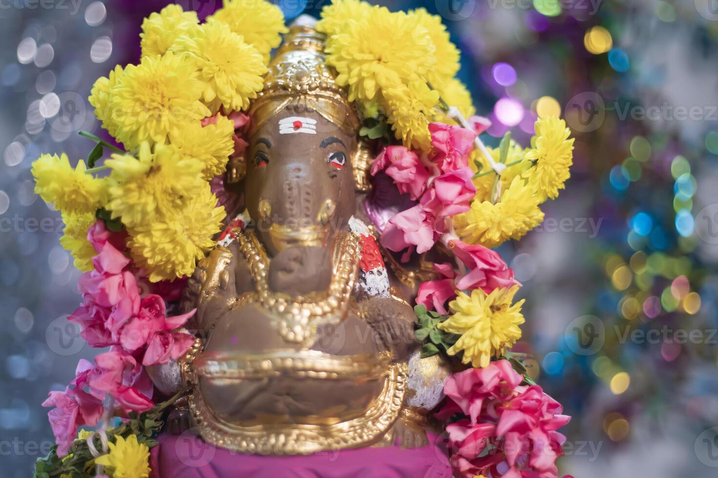 Hindu god Ganesha statue made of clay decorated with flower garlands on bokeh effect background. Closeup photo
