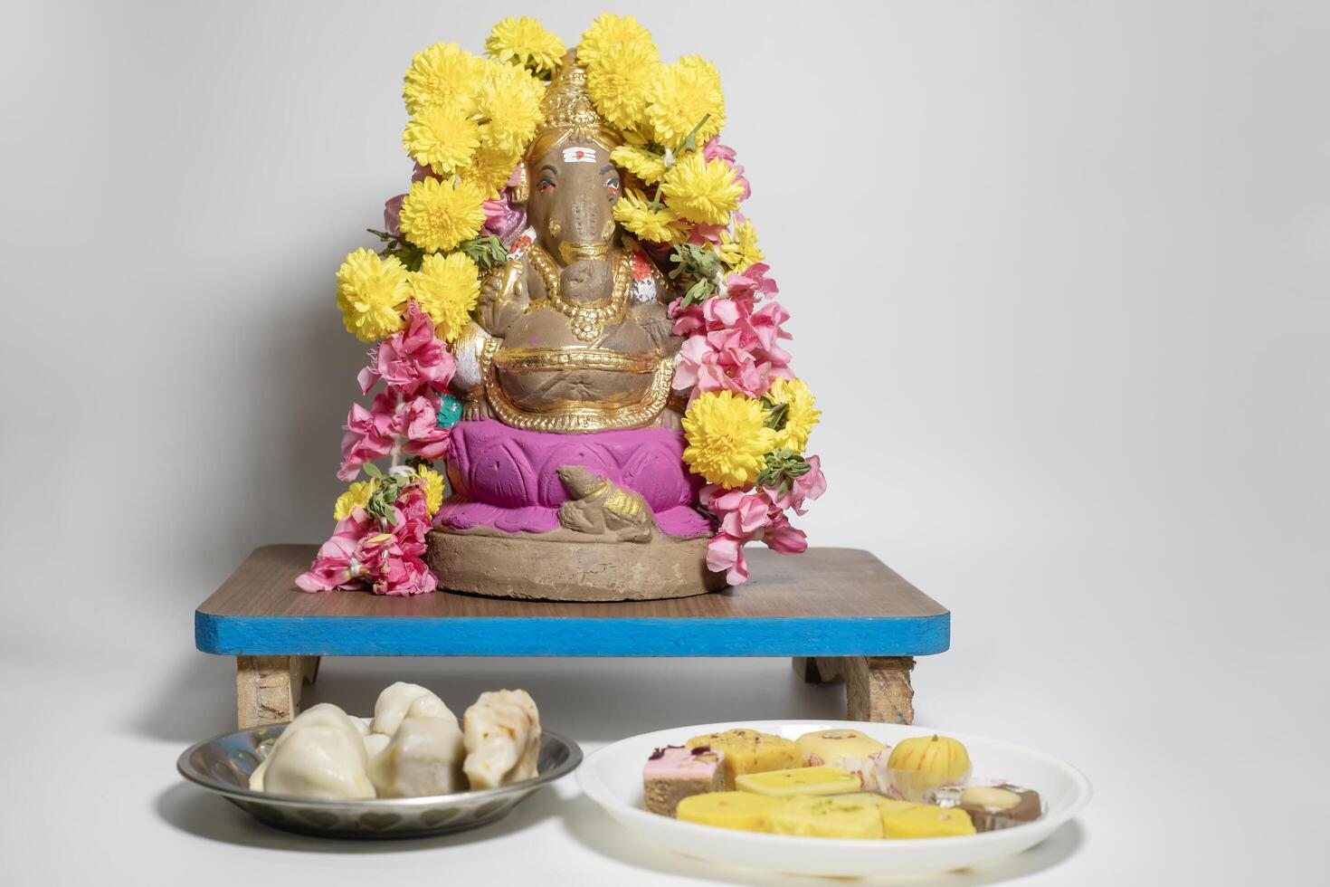 Ganesha idol made of clay decorated with flower garlands on wooden table with sweets on white background. Closeup photo