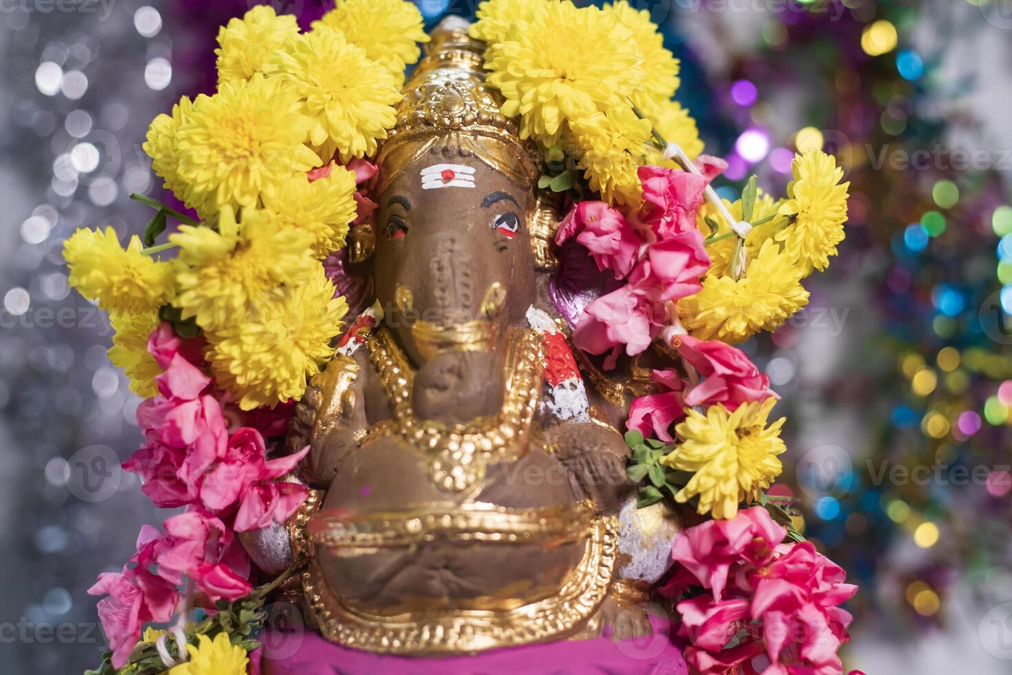 Hindu god Ganesha statue made of clay decorated with flower garlands on bokeh effect background. Closeup photo