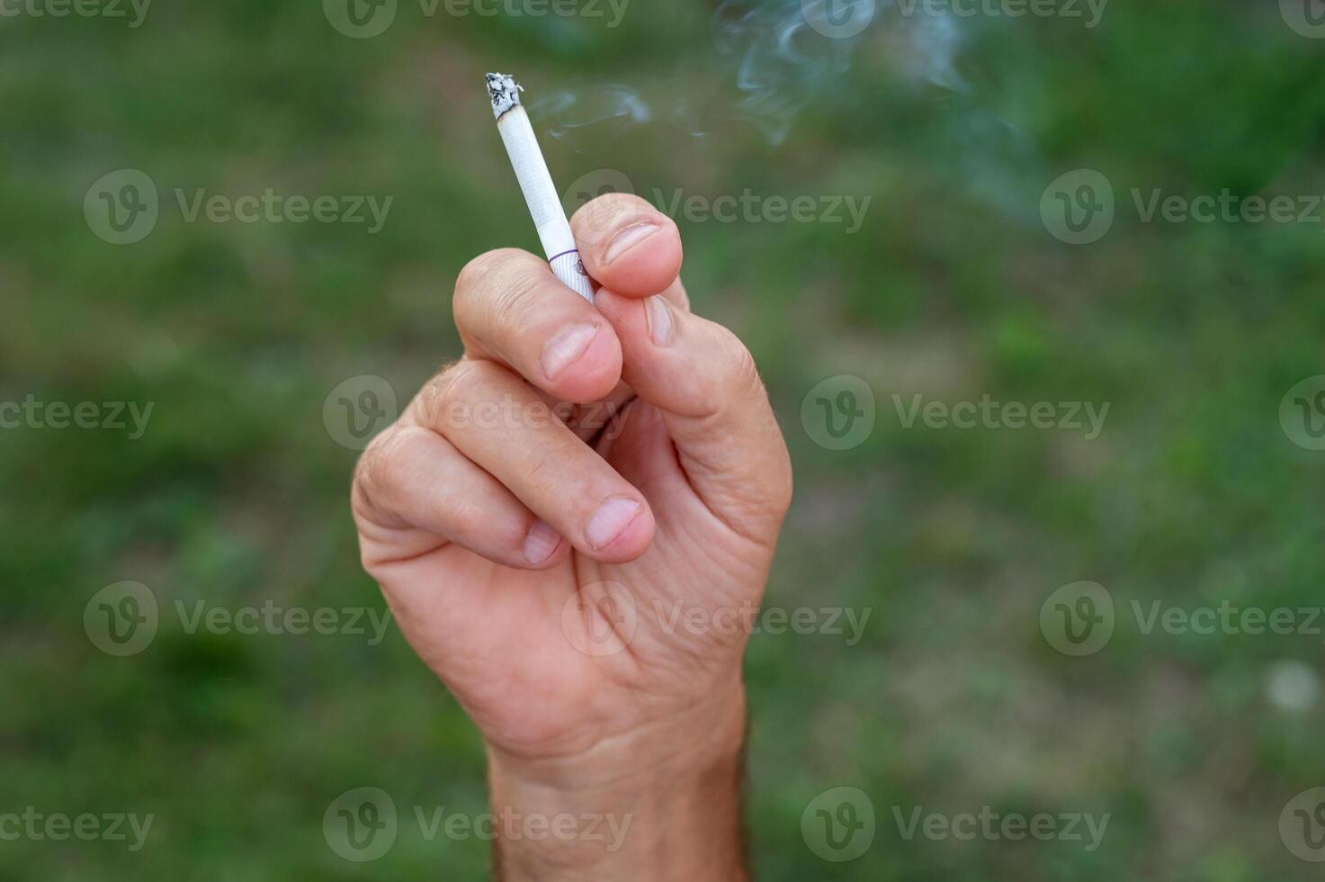 A man's hand holds a cigarette in his hand against a background of green grass. photo