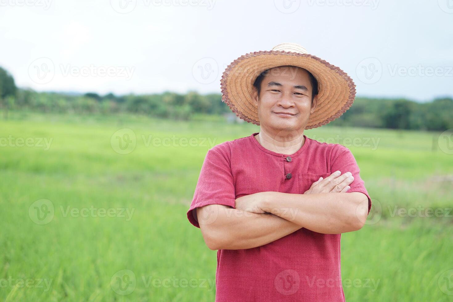 Handsome Asian man farmer is at paddy field, wears hat, red shirt, cross arms on chest, feels confident. Concept ,Agriculture occupation. Thai farmer. photo