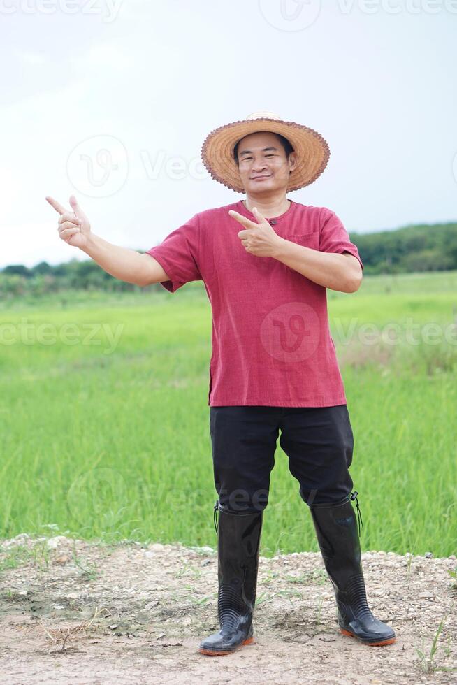 Handsome Asian man farmer stands at agricultural land, wears hat, makes hands gesture to point, feels confident. Full body shot. Concept, agriculture occupation. Thai farmer. Rural scene. photo