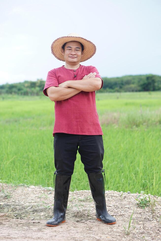 Handsome Asian man farmer stands at agricultural land, wears hat, cross arms on chest, feels confident. Full body shot. Concept, agriculture occupation. Thai farmer. Rural scene. photo
