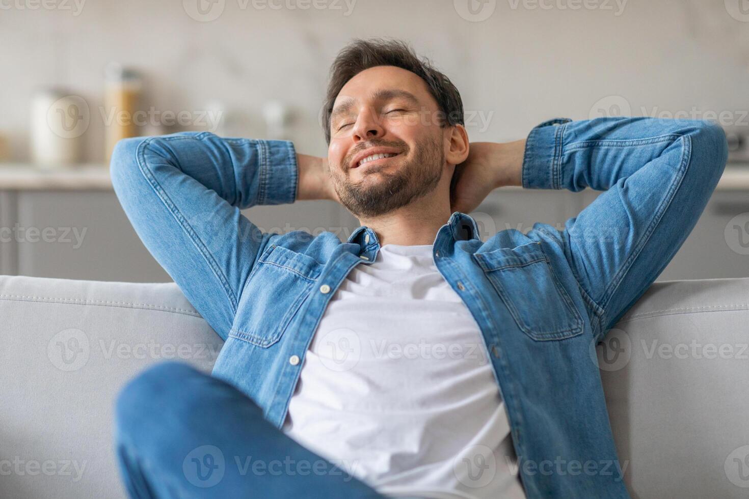 A man is sitting on a couch with his eyes closed. He is wearing a white t-shirt and a blue denim shirt. He has his hands behind his head and is leaning back on the couch. photo