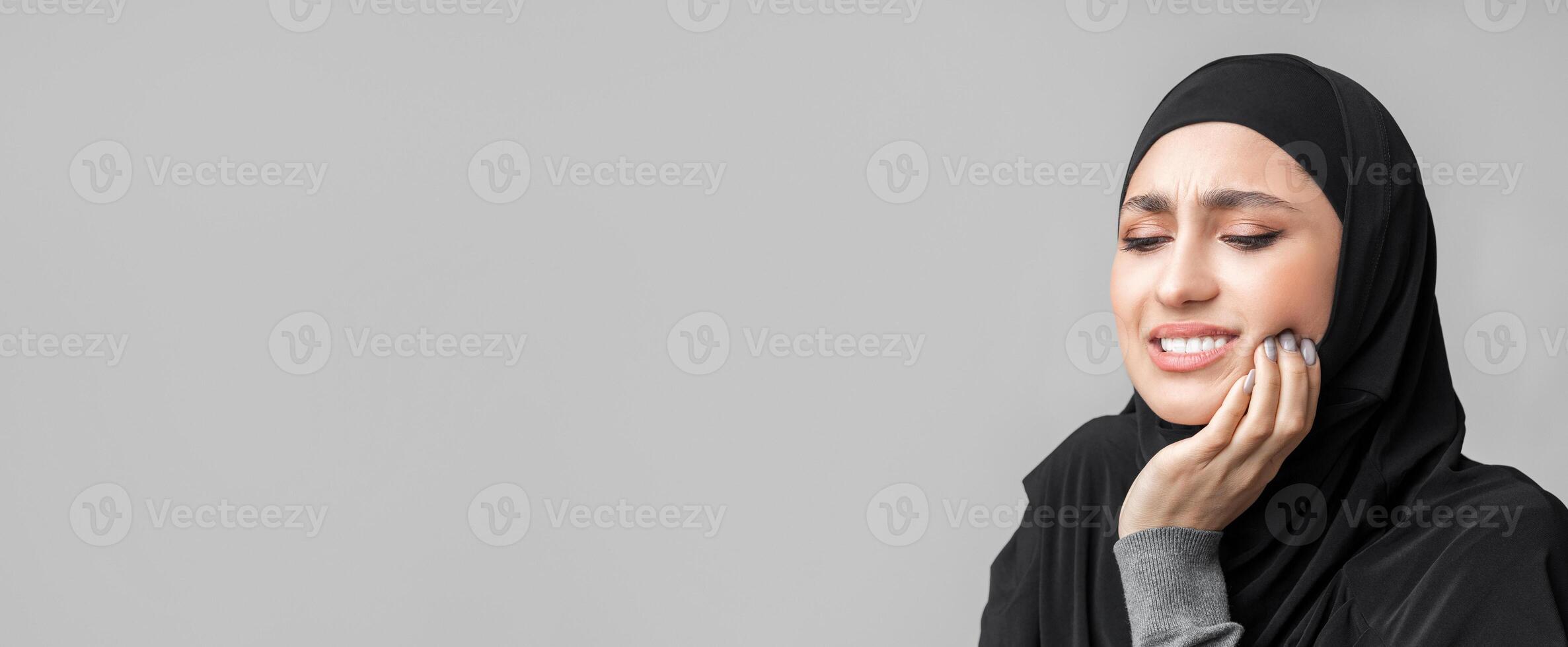 Tooth sensitivity. Young muslim woman in hijab suffering from teeth pain and touching her inflamed cheek, gray background with empty space, closeup photo