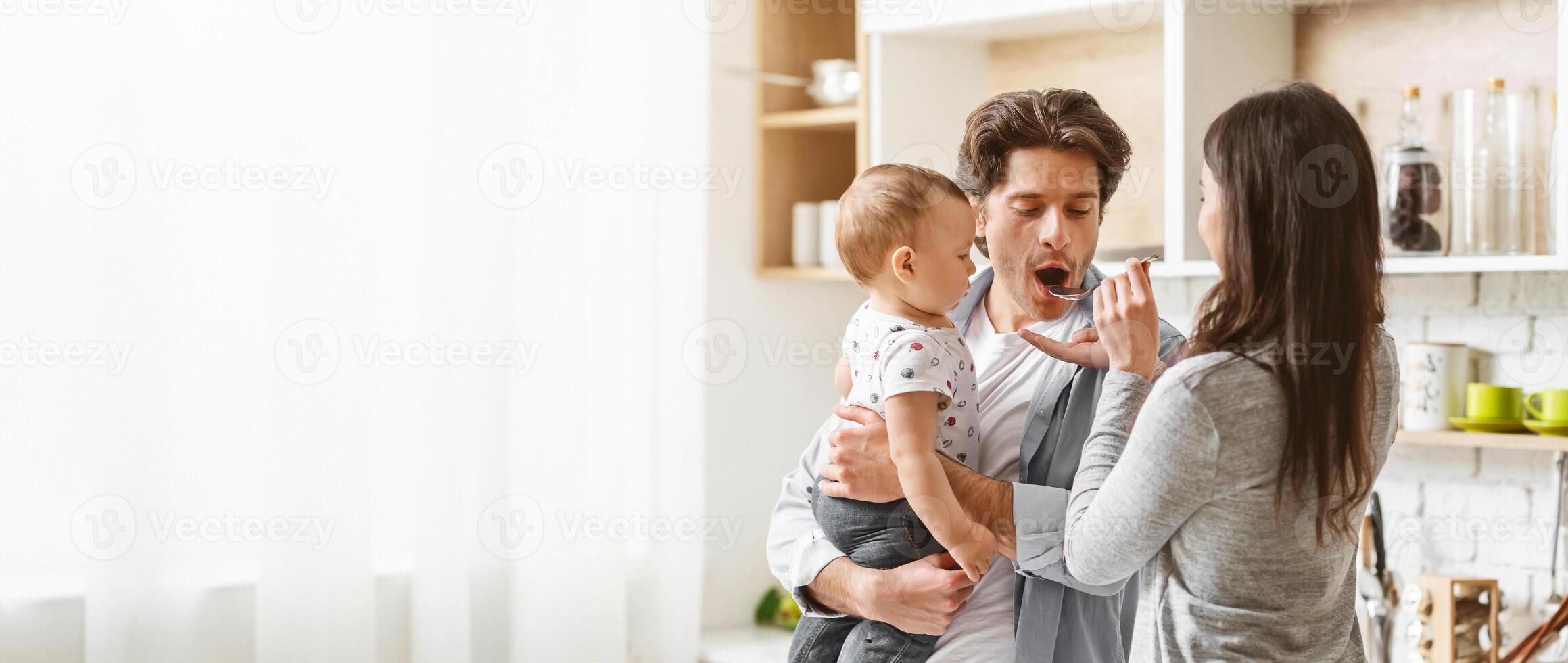 A father is feeding his baby with a spoon in the kitchen. He is holding the baby in his arms, while the baby is looking at the spoon and smiling, copy space photo