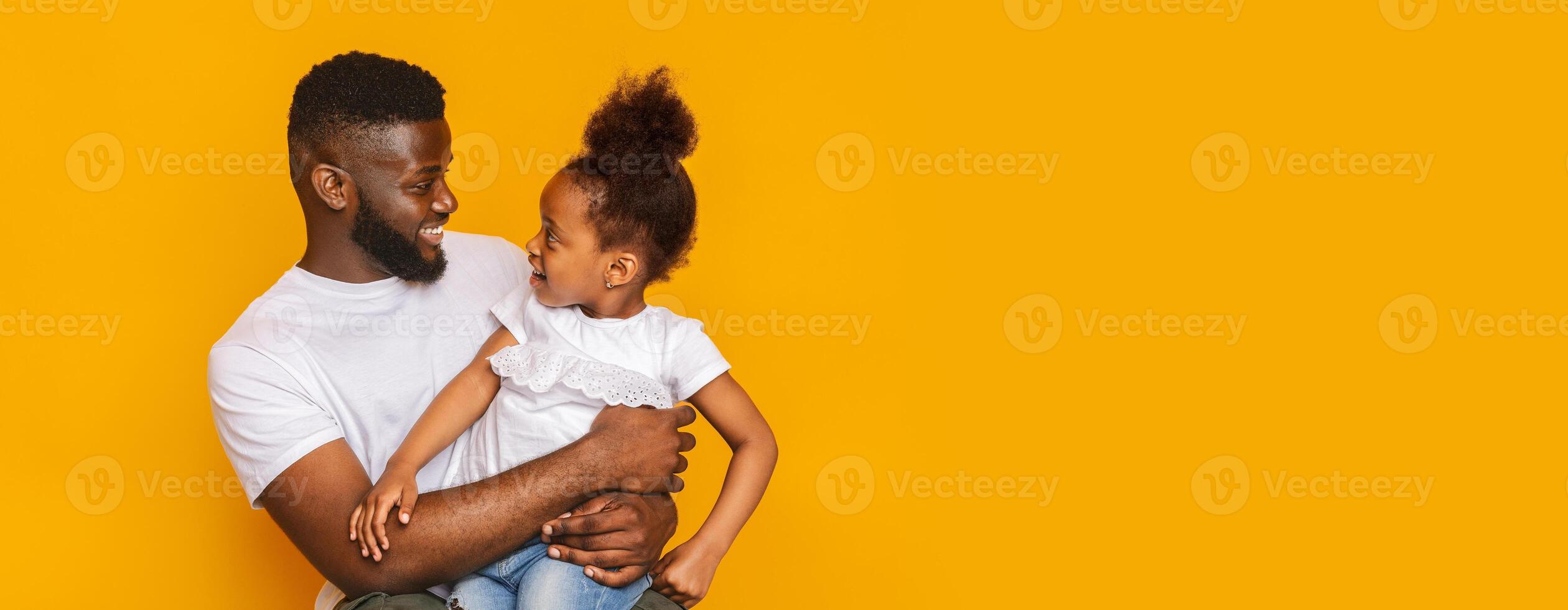 A joyful black father and his young daughter share a playful moment as they gaze into each other's eyes. Both are wearing casual, comfortable attire, and the bright yellow background photo