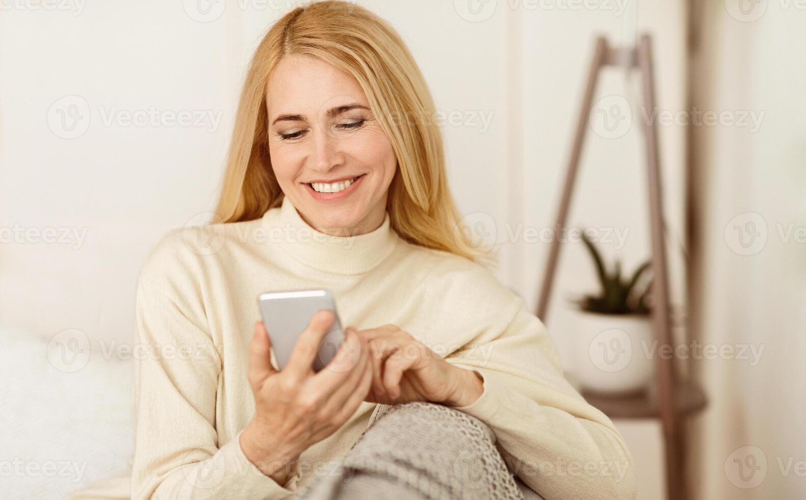 contento de edad mediana mujer surf Internet en teléfono, descansando en sofá a hogar foto