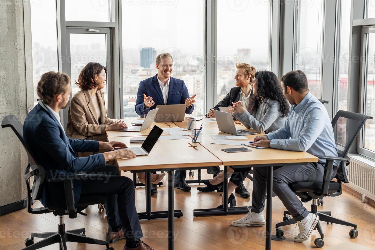A group of five professionals are seated around a large, rectangular table in a modern office. They are engaged in a discussion, looking at each other and using laptops photo