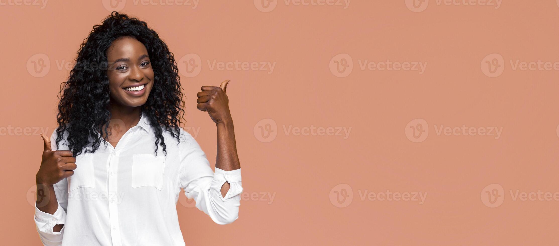 Smiling woman with curly black hair wearing a white button-down shirt. She is giving a thumbs-up gesture with her left hand and pointing with her right hand, standing in front of a peach background. photo