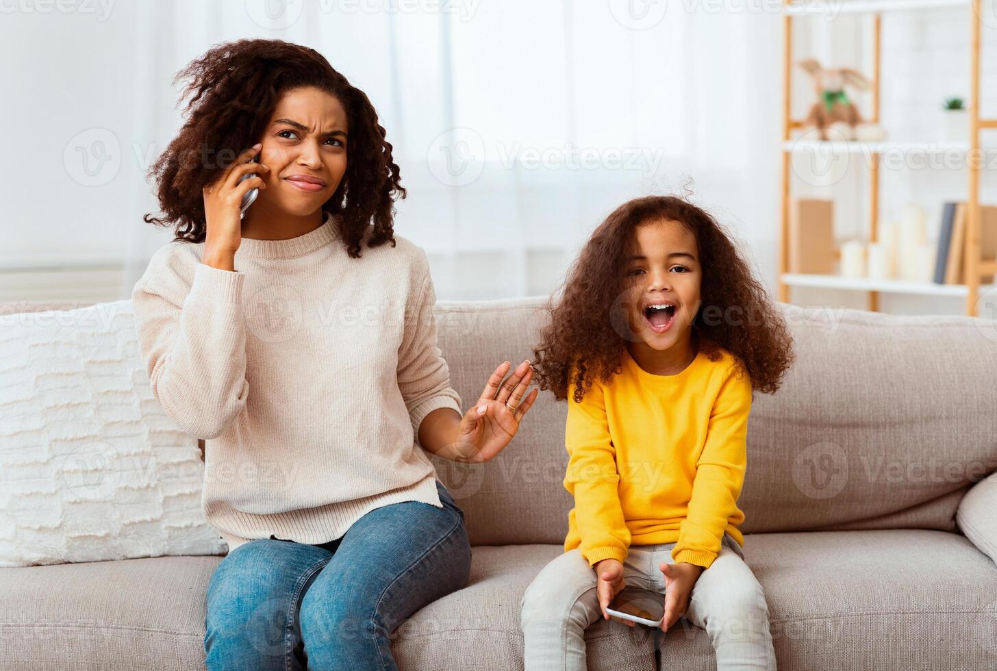 Loud Child. Little Daughter Shouting While Afro Mother Talking On Phone Sitting On Couch At Home photo