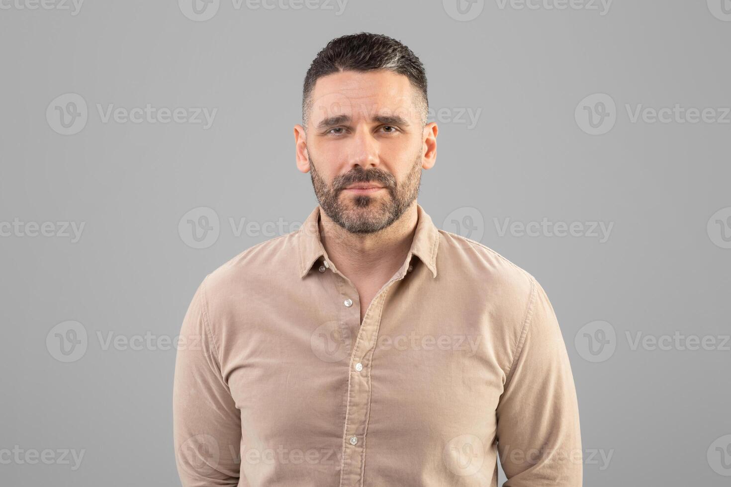 A man with a short haircut and beard is standing against a plain gray background, wearing a beige shirt and looking directly at the camera. The setting appears to be indoors with even lighting. photo