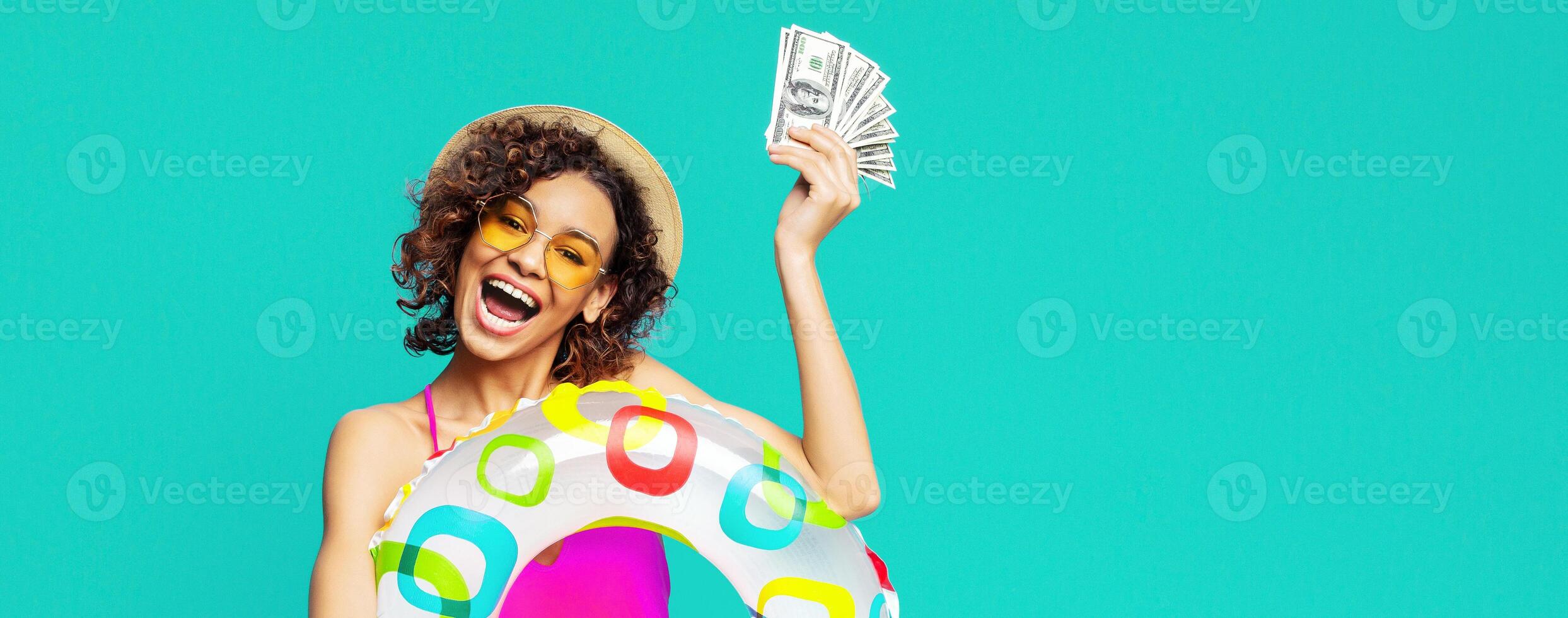 A young black woman in a swimsuit and a straw hat is holding a fan of cash in her right hand and a colorful inflatable ring in her left. The background is a solid, bright blue color. photo