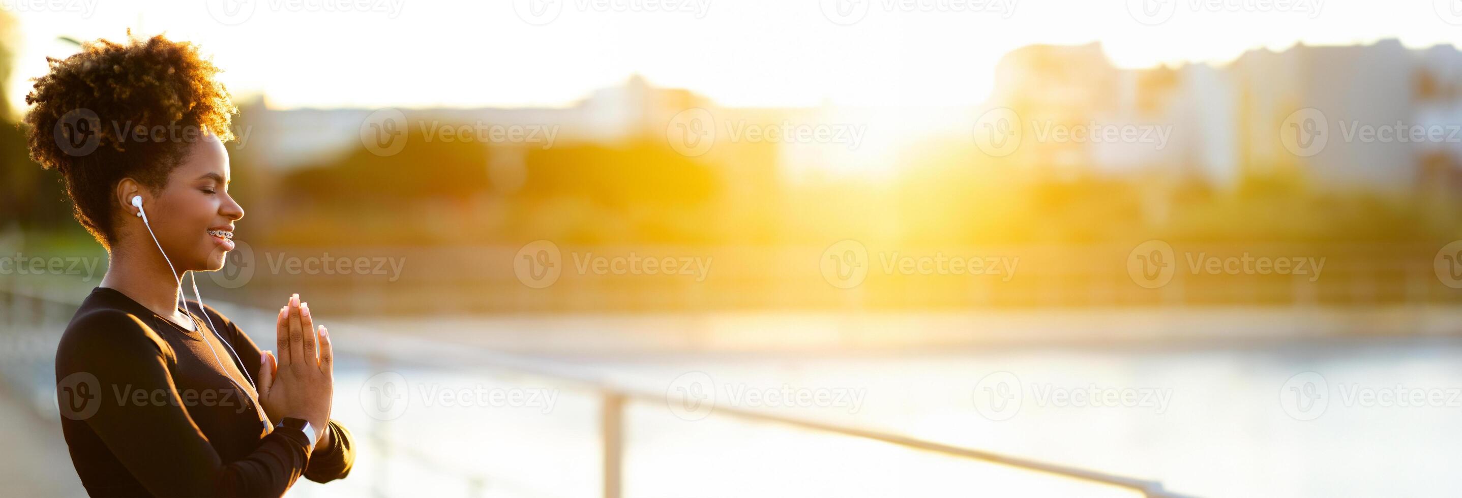 Yoga Outdoors. Portrait Of Smiling Young Black Woman Meditating Outside In The Morning, Sporty Lady Making Namaste Gesture With Closed Eyes And Listening Music In Earphones, Copy Space photo