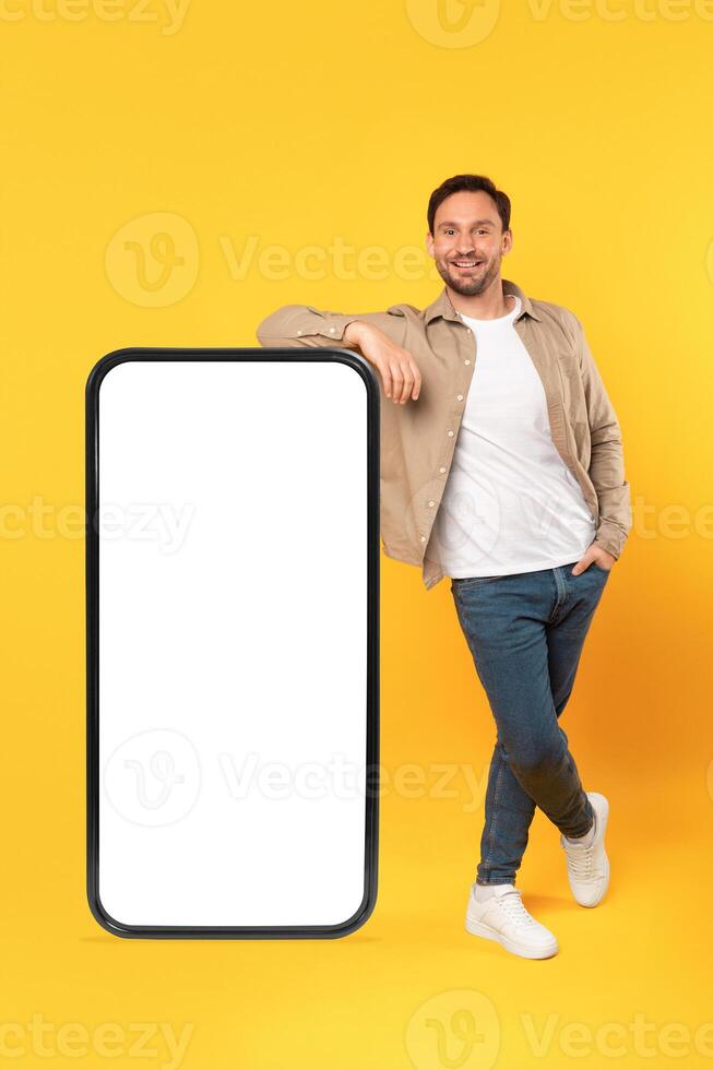 A smiling man in casual attire leans confidently against an oversized, blank white screen of a smartphone model, mockup copy space photo