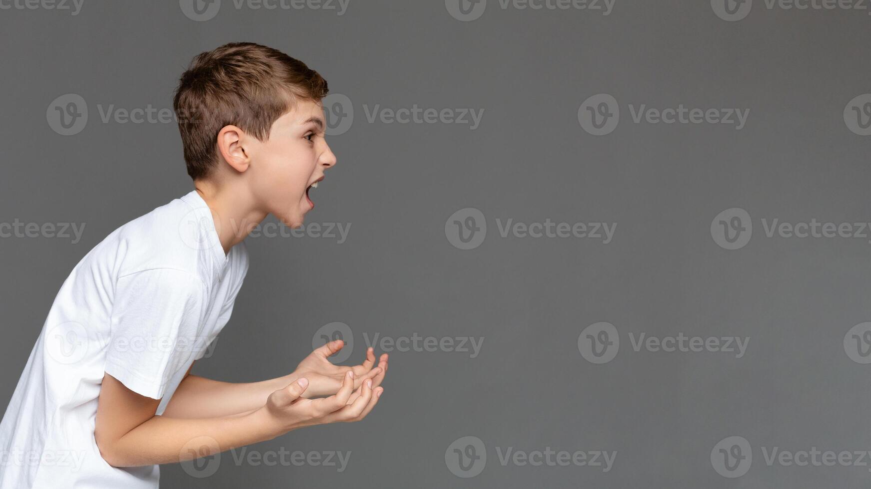 Awkward age. Angry emotional boy screaming and gesturing with hands in air, side view, gray studio background with free space photo