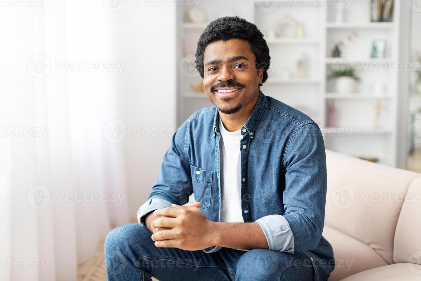 African American man is seated comfortably on a couch in a well-lit living room. The room is furnished with a coffee table, rug, and decorative items. photo