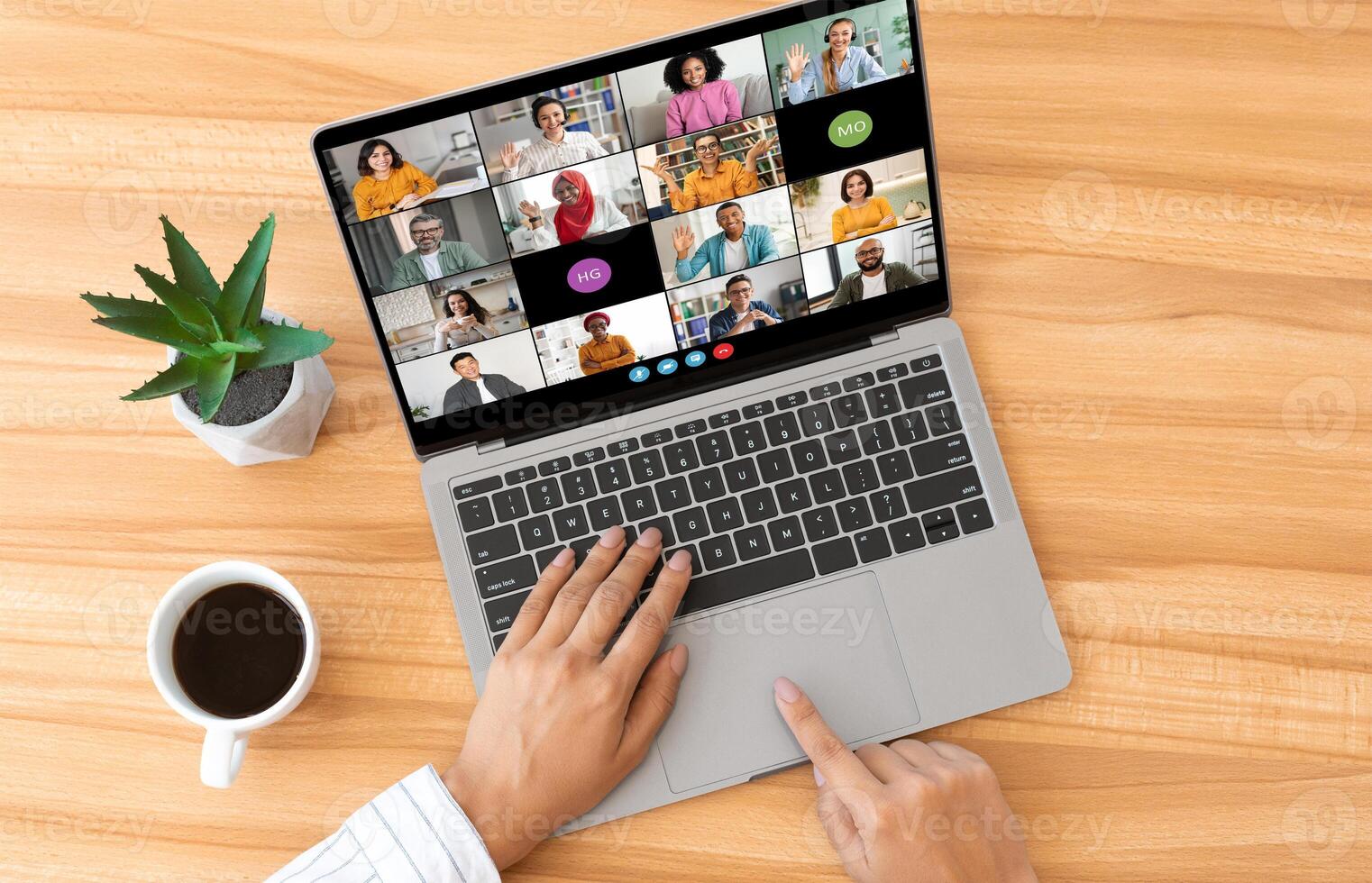 Businesswoman hands are shown typing on a laptop, which is open and displaying a conference call with multiple participants. The laptop is sitting on a wooden desk, top view photo