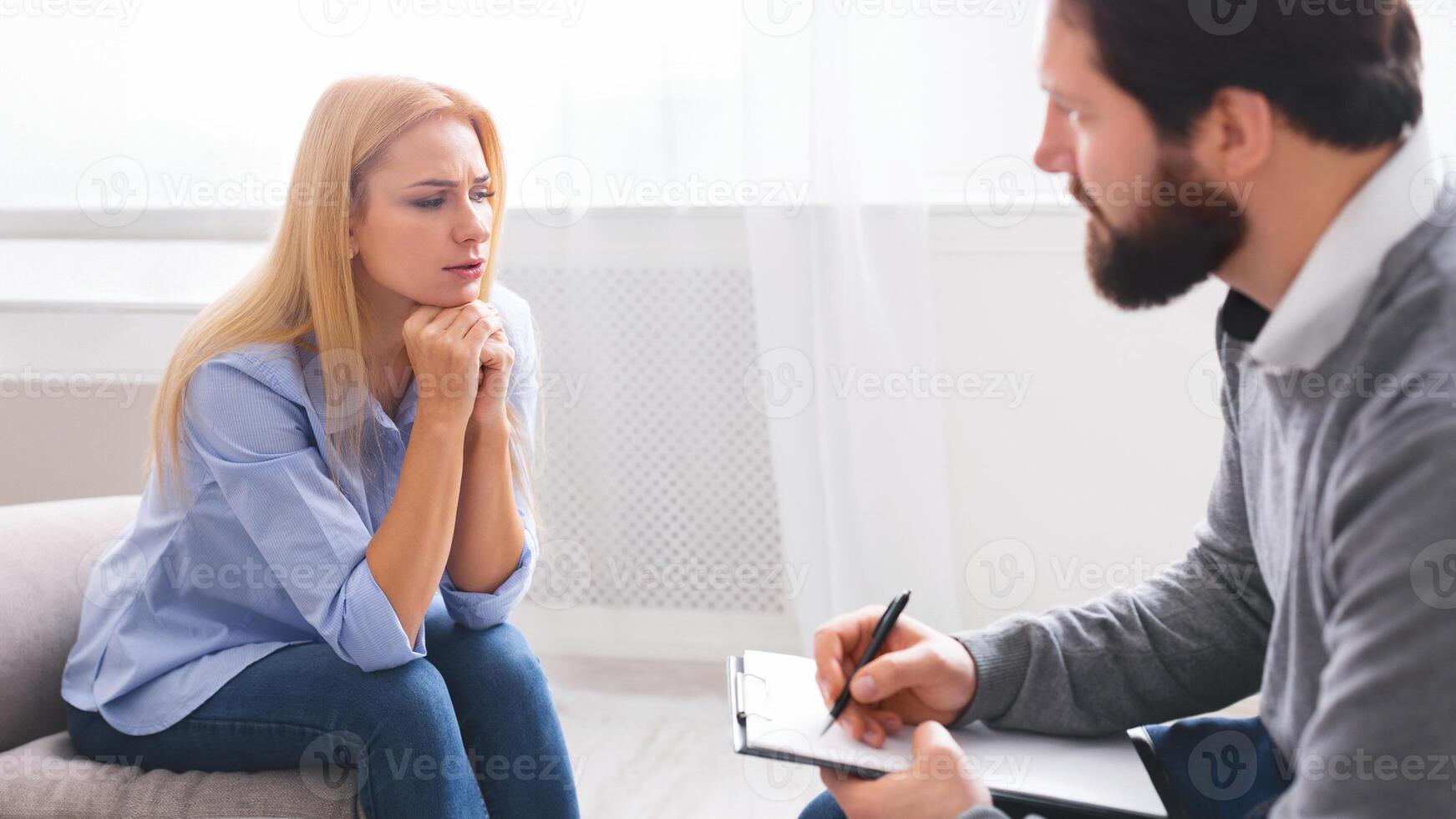 A thoughtful woman is engaged in a serious conversation with a man therapist who is taking notes. They are seated in a well-lit room with a neutral color scheme, indicating a professional setting photo