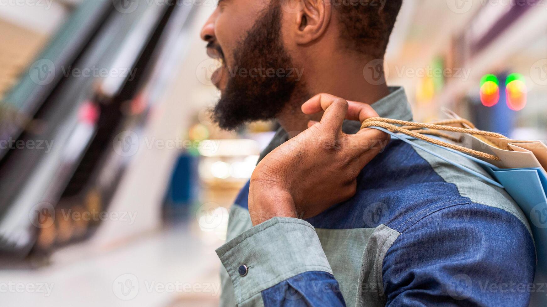 Closeup of black man is seen holding multiple shopping bags in a bustling mall, surrounded by various stores and people, focused on his purchases, navigating through the crowded shopping center. photo