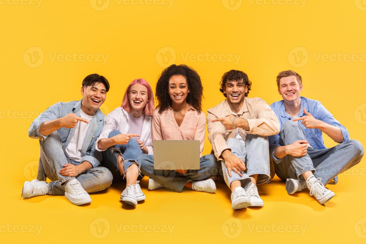 A diverse group of five young adults, all smiling and looking at the camera, are sitting on a yellow background. They are holding a laptop and pointing at it, as if showing it off to someone photo