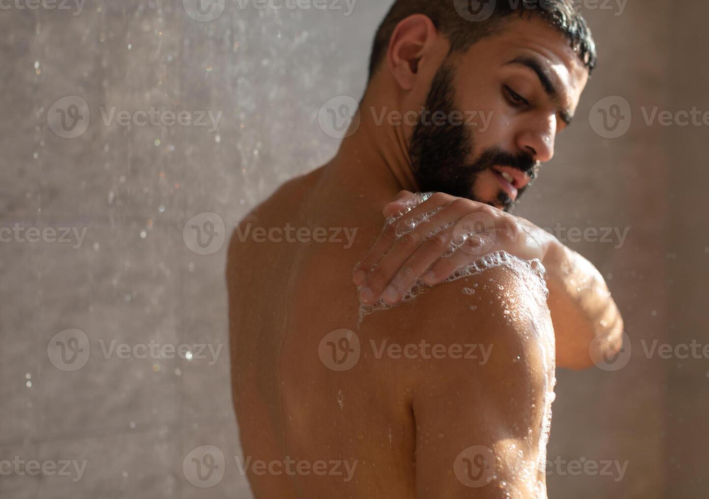 Skincare Concept. Young muscular man taking refreshing shower washing his body, back and bare shoulders with soap foam. Confident handsome guy standing in the bathroom under falling hot water drops photo