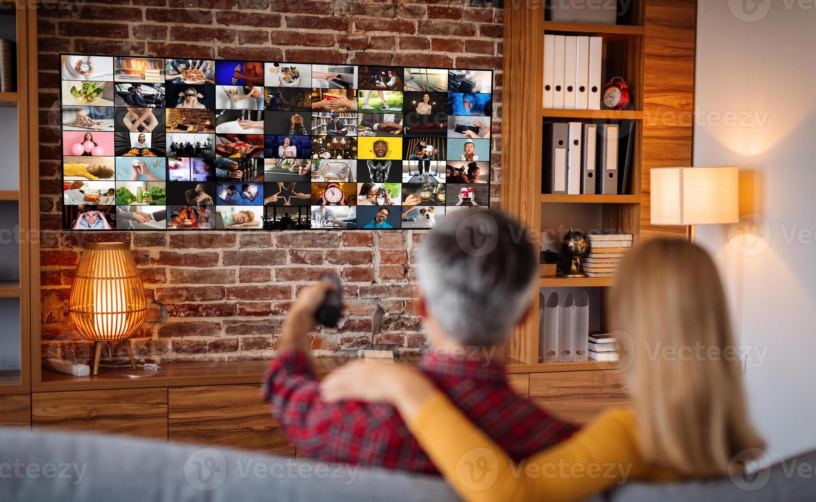 A couple sits comfortably on a couch in a living room, watching television. The television screen displays a grid of various images, likely from different channels or streaming services. photo