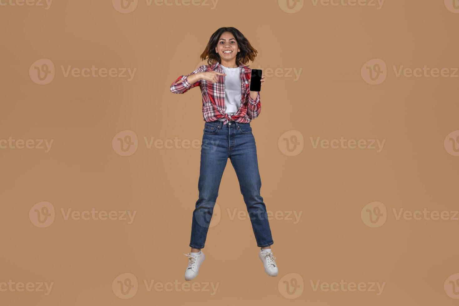 A young middle eastern woman with long brown hair is jumping in the air while smiling and holding a smartphone in her right hand. The background is a solid beige color. photo
