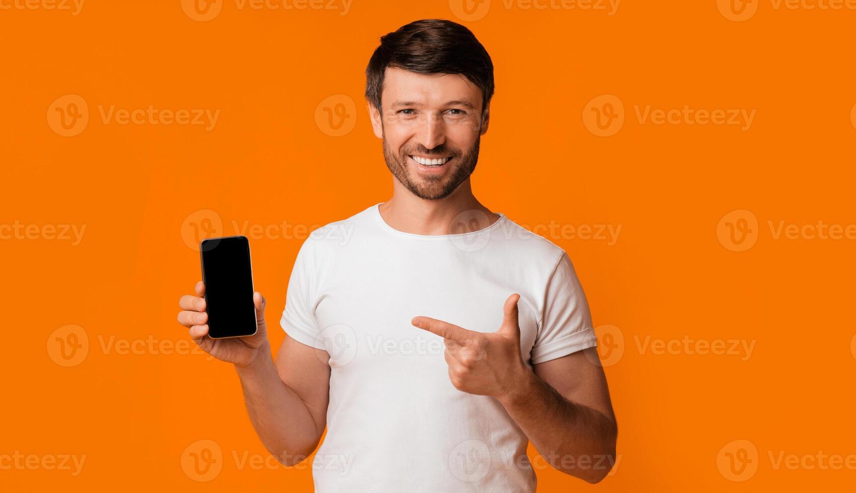 A man in a white t-shirt holds a black smartphone with a blank screen and points to it with a smile, standing against an orange background. photo