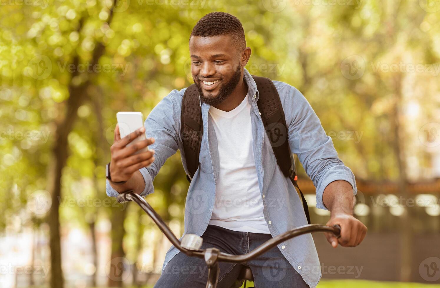 Cheerful African American Guy Having Call While Driving Bicycle Outdoors, Holding Smartphone And Smiling, Selective Focus photo