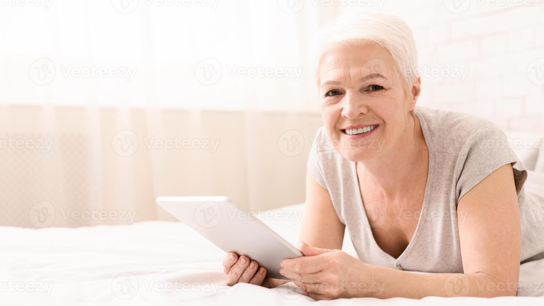 Elderly woman is reclining on a bed while holding a tablet in her hands. She appears engrossed in the devices screen, possibly reading or watching something, copy space photo