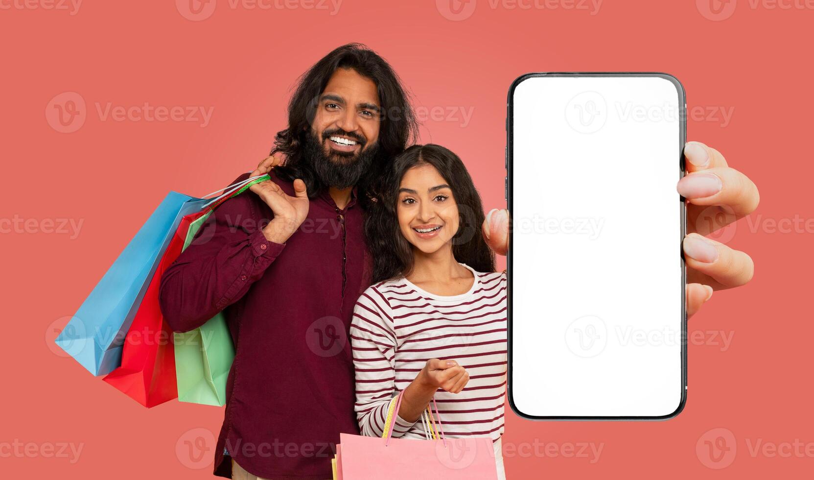 A smiling Indian couple stands in front of a pink background, holding shopping bags and a large smartphone with a blank screen. photo
