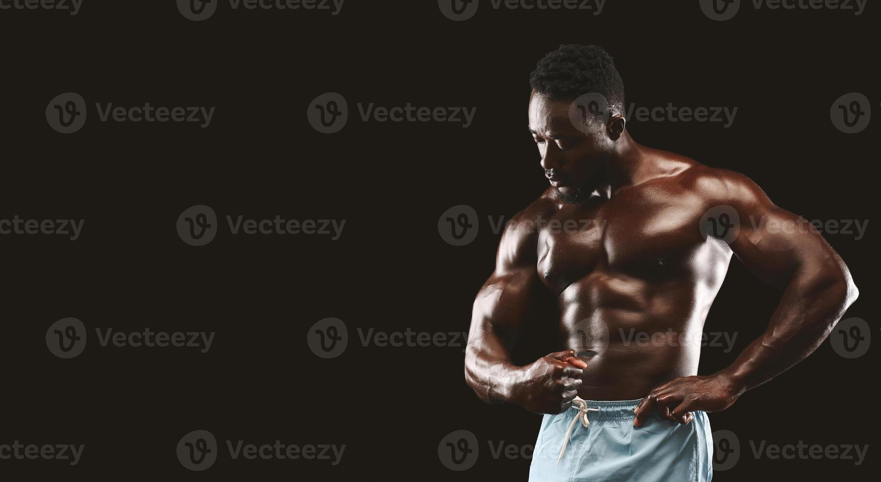 A muscular man poses in front of a black background, showing off his toned physique. He is wearing blue athletic shorts and has a serious expression on his face. photo